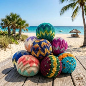 Puede incluir: Una colección de coloridos sacos de hacky hechos a mano en varios patrones y tonos, incluyendo azul, verde, rosa y naranja. Las bolas están apiladas sobre una superficie de madera con una playa y un océano de fondo, y palmeras.