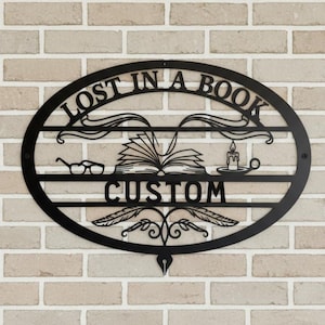 Könnte beinhalten: Schwarze Metall-Wandkunst mit dem Text "LOST IN A BOOK CUSTOM" in einem ovalen Rahmen. Das Design umfasst ein offenes Buch, eine Brille, eine Kerze und dekorative Elemente, montiert an einer Ziegelmauer.