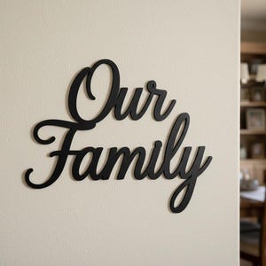 Könnte beinhalten: Schwarze Wandkunst aus Holz mit dem Schriftzug "Our Family" in einer Schreibschrift. Das Schild ist an einer cremefarbenen Wand angebracht. Der Hintergrund zeigt einen Einblick in ein Esszimmer mit einem Holztisch und Stühlen.