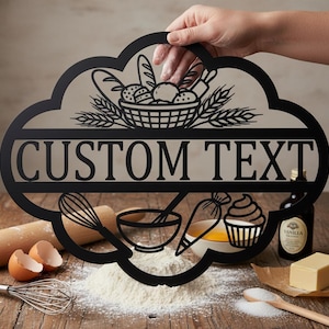 Könnte beinhalten: Schwarzes Metallschild in Wolkenform mit den Worten "CUSTOM TEXT" und Backmotiven. Das Schild zeigt einen Brotkorb, Weizen, einen Schneebesen, eine Schüssel, einen Spritzbeutel, einen Cupcake und andere Backutensilien.