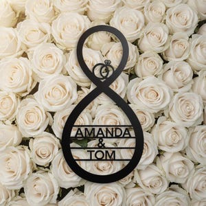 Op de afbeelding: Zwart metalen trouwteken met de namen "AMANDA & TOM" en twee in elkaar grijpende ringen met een diamant. Het teken heeft de vorm van een oneindigheidssymbool, tegen een achtergrond van witte rozen.