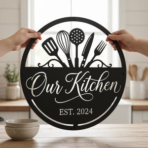 Könnte beinhalten: Schwarzes Küchenschild aus Metall mit kreisförmigem Design. Das Schild zeigt ausgeschnittene Silhouetten von Küchenutensilien über den Worten "Our Kitchen" und "EST. 2024". Das Schild wird von zwei Händen gehalten.