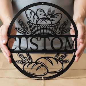 Könnte beinhalten: Schwarzes Metallschild mit einem kreisförmigen Design mit einem Brotkorb, Weizenähren und dem Wort "CUSTOM". Das Schild hat ein dekoratives Design von zwei Brotlaiben am unteren Rand. Das Schild wird von einer Person gehalten.