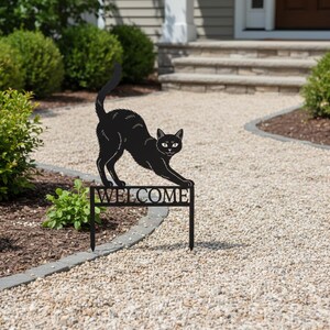 Könnte beinhalten: Ein schwarzer Metall-Katzen-Gartenstecker mit dem ausgeschnittenen Wort "WELCOME" unter der Katze. Die Katze befindet sich in einer Dehnpose. Der Stecker befindet sich in einem Kiesweg in der Nähe eines Gartens.