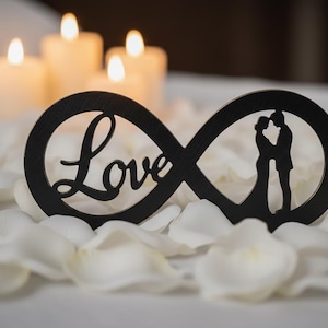Könnte beinhalten: Ein schwarzes Holz-Unendlichkeitszeichen mit dem Wort "Love" und einer Silhouette eines sich umarmenden Paares. Das Symbol liegt auf weißen Blütenblättern, mit brennenden Kerzen im Hintergrund.