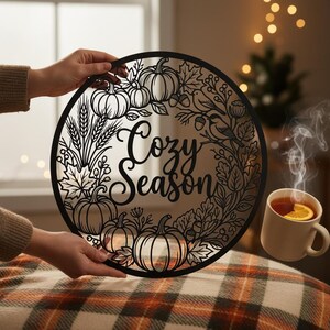 Könnte beinhalten: Ein kreisförmiges Wandkunstobjekt aus schwarzem Metall mit dem Schriftzug "Cozy Season" in der Mitte, umgeben von einem detaillierten Scherenschnitt-Design aus Kürbissen, Weizenähren, Ahornblättern, Eicheln und kleinen Vögeln.
