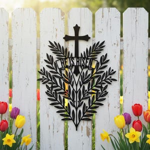 Könnte beinhalten: Schwarze Metall-Osterdekoration mit einem Kreuz über einem Banner mit der Aufschrift "HE IS RISEN" und einem Blätterdesign. Die Dekoration ist an einem weißen Lattenzaun befestigt, mit bunten Tulpen und Narzissen im Vordergrund.