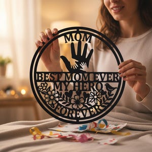 Puede incluir: Arte de pared de metal negro con las palabras "MOM", "BEST MOM EVER", "MY HERO" y "FOREVER". El diseño incluye una mano que sostiene una mano más pequeña, flores y un pájaro. Un regalo reflexivo.