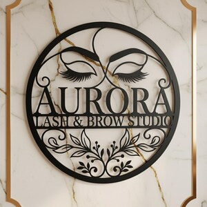 Könnte beinhalten: Schwarzes Metallschild mit der Aufschrift "AURORA LASH & BROW STUDIO". Das runde Design zeigt stilisierte Wimpern und florale Elemente. Das Schild ist an einer Wand mit Marmormuster befestigt.