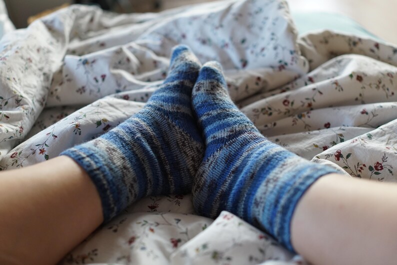 Pode incluir: Close-up de dois p&eacute;s usando meias listradas azuis e brancas. As meias est&atilde;o em uma cama com uma colcha estampada floral. As meias t&ecirc;m uma apar&ecirc;ncia texturizada.