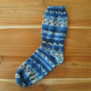 Pode incluir: Uma meia de malha azul e branca com um padr&atilde;o listrado. A meia apresenta v&aacute;rios tons de azul e branco, com um design texturizado. A meia &eacute; exibida em uma superf&iacute;cie de madeira.