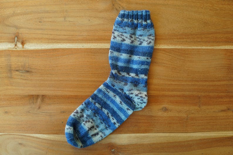 Pode incluir: Uma meia de malha azul e branca com um padr&atilde;o listrado. A meia apresenta v&aacute;rios tons de azul e branco, com um design texturizado. A meia &eacute; exibida em uma superf&iacute;cie de madeira.