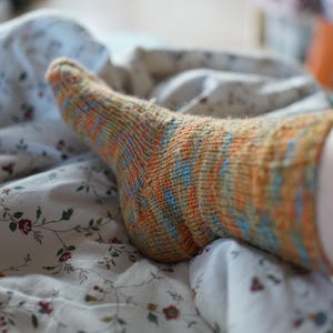 Pode incluir: Close-up de uma meia de malha com listras laranja, azuis e amarelas. A meia est&aacute; em uma cama com um len&ccedil;ol estampado floral. A meia parece ser feita &agrave; m&atilde;o.