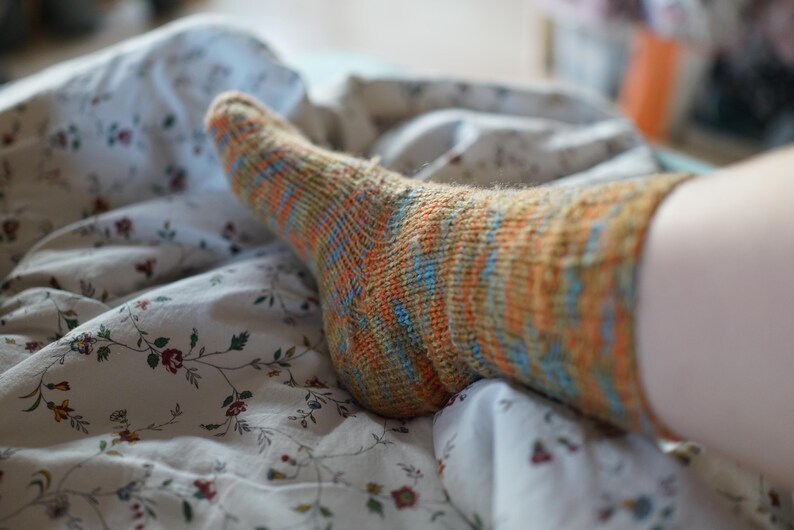 Pode incluir: Close-up de uma meia de malha com listras laranja, azuis e amarelas. A meia est&aacute; em uma cama com um len&ccedil;ol estampado floral. A meia parece ser feita &agrave; m&atilde;o.