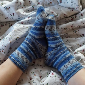 May include: A pair of blue and white striped knitted socks. The socks are resting on a floral patterned bedsheet. The socks have a mix of blue shades and white stripes, with a textured appearance. The bedsheet has a white background with small red and blue floral patterns.