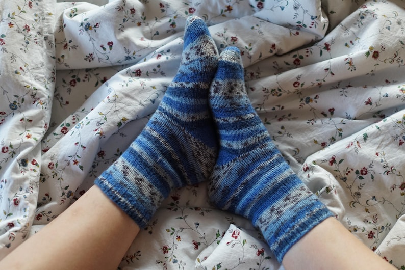 Pode incluir: Um par de meias de malha listradas em azul e branco. As meias est&atilde;o sobre um len&ccedil;ol com estampa floral. As meias t&ecirc;m uma mistura de tons de azul e listras brancas, com uma apar&ecirc;ncia texturizada. O len&ccedil;ol tem um fundo branco com pequenos padr&otilde;es florais vermelhos e azuis.