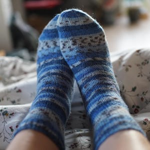 Pode incluir: Close-up de duas meias azuis de malha com um padr&atilde;o de riscas e pequenas formas geom&eacute;tricas. As meias est&atilde;o numa cama coberta com um len&ccedil;ol com padr&atilde;o floral. As meias s&atilde;o em tons de azul, cinzento e castanho.