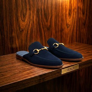 Peut inclure: Une paire de mules en daim bleu marine dotée d'un détail mors en métal doré sur le dessus. Ces chaussures à enfiler présentent un petit talon marron et un bout arrondi, présentées comme une paire de chaussures sur une étagère en bois.