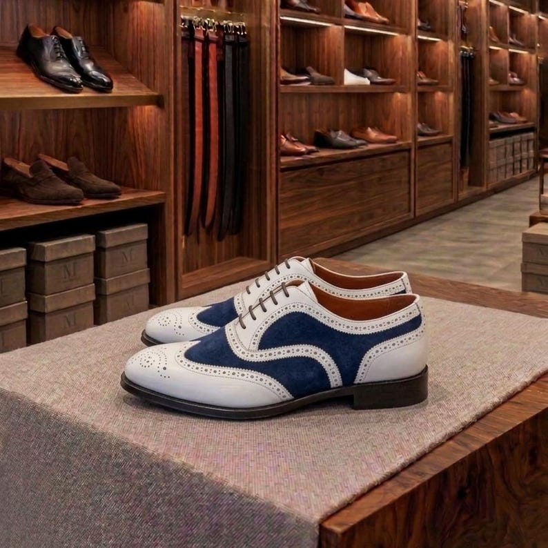 May include: A pair of two-tone oxford shoes featuring white leather and navy blue suede panels with decorative brogue detailing, thin brown laces, and a low stacked heel, displayed on a textured surface in a retail environment.