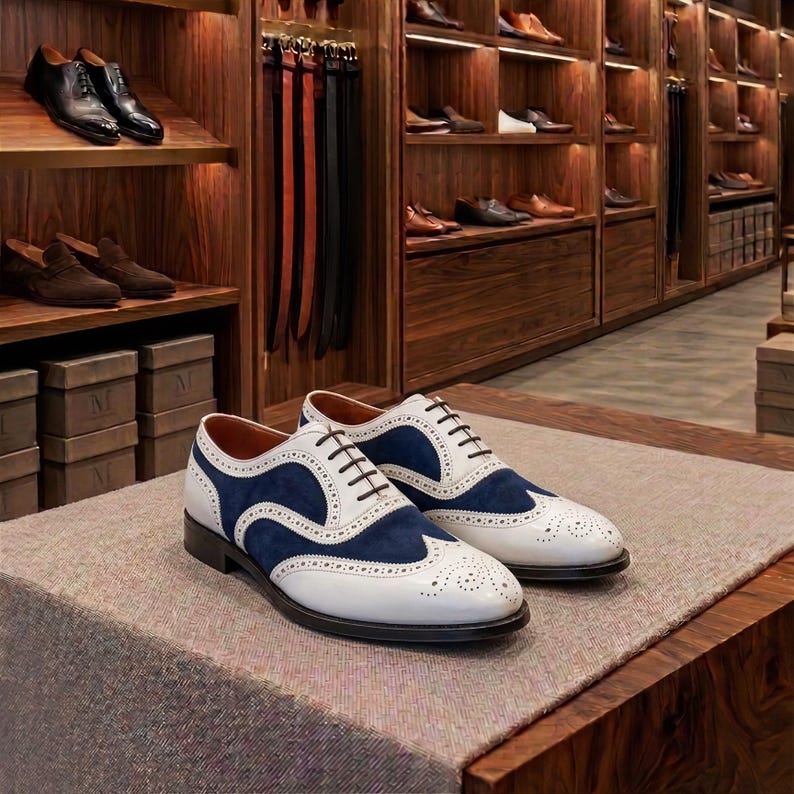 May include: A pair of two-tone oxford shoes featuring white leather and navy blue suede panels with decorative brogue detailing, a wingtip design, and dark brown laces, displayed on a textured surface in a retail store setting.