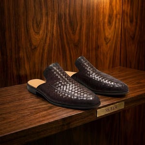 May include: A pair of dark brown mules featuring a woven leather upper with a pointed toe and a low heel. The interior footbed is tan, and a small gold-toned metal plaque with the text MULES is positioned on the edge of the wooden shelf.