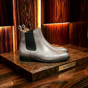 Könnte beinhalten: Ein Paar hellgraue Chelsea-Stiefel aus Leder mit schwarzen elastischen Seiteneinsätzen, einer Zugschlaufe an der Ferse und einem niedrigen schwarzen Absatz. Die Stiefel stehen auf einem Holzblock mit einem Metallschild, auf dem BOOTS steht.