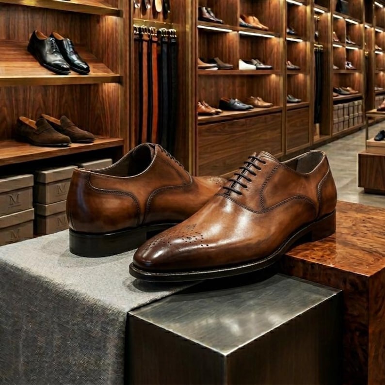 May include: A pair of brown leather dress shoes with intricate brogue detailing on the toe. The shoes have a classic lace-up design and are displayed on a textured surface. The background features a wooden shoe display.
