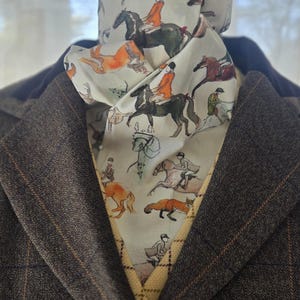 以下が含まれることがあります： クリーム色の、馬に乗ったライダー、キツネ、犬の水彩画プリントが施されたアスコットスカーフ。 スカーフは、微妙な格子柄と黄色の模様の裏地が付いた茶色のツイードジャケットの上にドレープされています。
