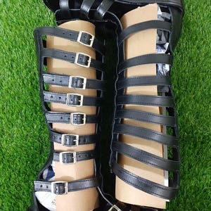 May include: Black gladiator sandals with multiple straps and silver buckles. The sandals have a platform sole and extend to the knee. The sandals are displayed on a green surface.