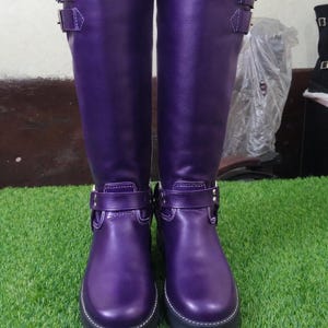 May include: A pair of knee-high purple boots with silver buckles and white stitching. The boots have a rounded toe and a thick black sole. The boots are made of a smooth, shiny material.