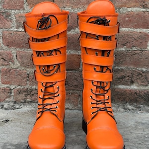 May include: A pair of bright orange mid-calf boots featuring black laces and four horizontal straps with silver-toned buckles across the front. The boots have a rounded toe, a thick black lug sole with white stitching, and a side zipper closure.
