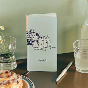 以下が含まれることがあります： 漫画のキャラクターのイラストが描かれた、水色の2026年版手帳。表紙には、キャラクターのグループと吹き出しの中のテキストが描かれています。手帳の横にはペンと水が入ったグラスがあります。