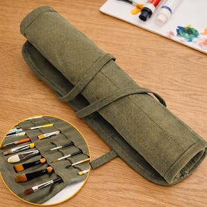 May include: An olive green canvas brush roll, partially unrolled, displaying various paint brushes. The roll is secured with a matching fabric strap. A palette with paint tubes and a brush is visible in the background. The roll is designed for artists.