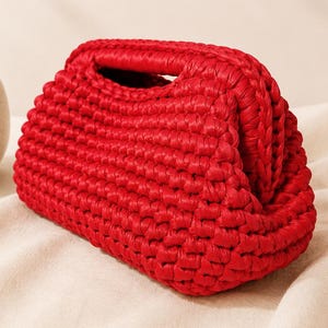 May include: A vibrant red clutch bag crafted with a thick, textured crochet knit pattern. The bag features a structured, rounded frame with a top opening and a consistent, dense stitch throughout its exterior, showcasing a bold, solid color.