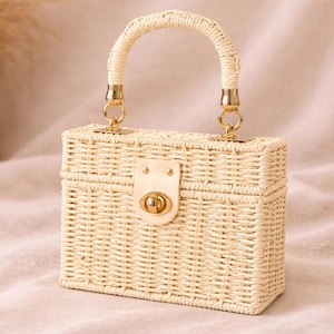 May include: A rectangular woven straw handbag with a curved handle and gold-colored hardware. The bag features a flap closure with a gold-colored clasp. The bag is a light beige color.