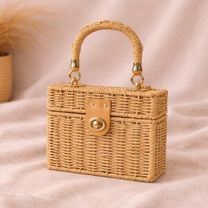May include: A rectangular woven handbag with a curved handle and gold-colored hardware. The bag features a tan leather closure with a gold-colored clasp. The bag is made of a light brown woven material.