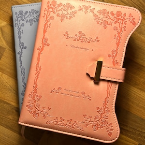 Könnte beinhalten: Zwei ledergebundene Notizbücher, eines hellblau und eines korallenrosa, mit geprägten Rosendesigns. Das rosa Notizbuch hat einen goldenen Verschluss und den Text "For the change that you wish to see in the world."