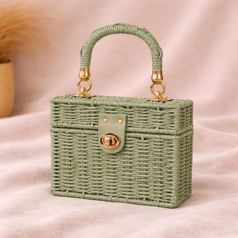 May include: A rectangular, sage green woven handbag with a curved handle and gold-colored hardware. The bag features a clasp closure and a structured design, suitable for carrying small items. The handle is also woven, matching the bag's texture.