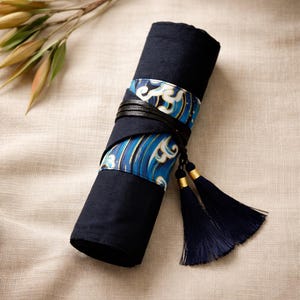 Op de afbeelding: Een opgerolde marineblauwe stoffen etui met een blauw-wit golvenpatroonband en een zwarte leren band. Marineblauwe kwasten met gouden accenten hangen aan de onderkant. De etui is waarschijnlijk voor het opbergen van kunstbenodigdheden.