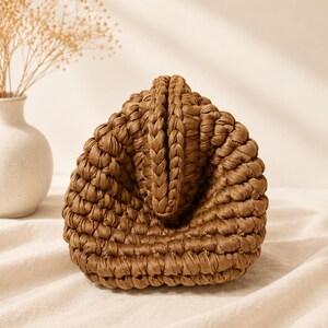 Pu&ograve; includere: Una borsa a mano marrone realizzata con un motivo a uncinetto intrecciato spesso. La borsa presenta una silhouette triangolare strutturata con una prominente cucitura centrale verticale e una base arrotondata, mostrando una costruzione in filato densa e strutturata.