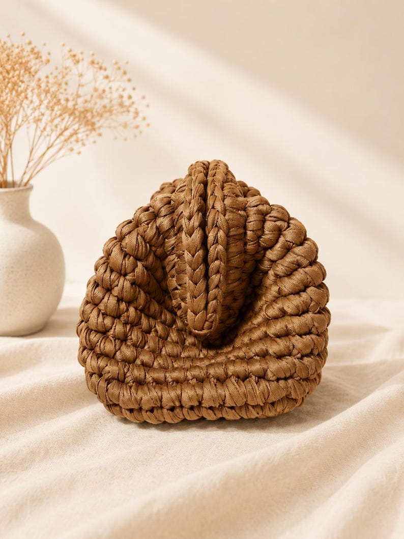 Pu&ograve; includere: Una borsa a mano marrone realizzata con un motivo a uncinetto intrecciato spesso. La borsa presenta una silhouette triangolare strutturata con una prominente cucitura centrale verticale e una base arrotondata, mostrando una costruzione in filato densa e strutturata.