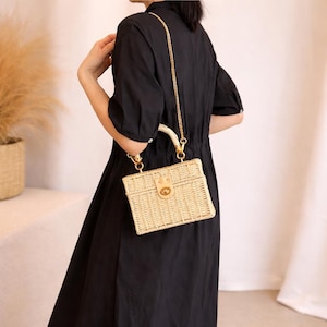 May include: A light beige woven handbag with a gold-tone chain strap and a small handle. The bag has a rectangular shape with a decorative clasp. The bag is carried by a person wearing a black dress.