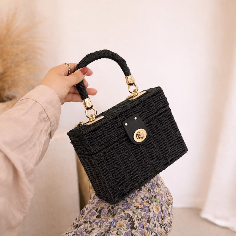 May include: A black woven handbag with a structured rectangular shape. The bag features a curved handle with gold-colored accents and a matching clasp. The bag is held by a person.
