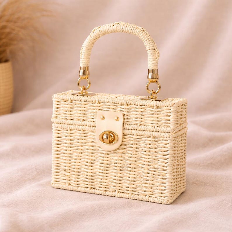May include: A rectangular woven straw handbag with a curved handle and gold-colored hardware. The bag features a flap closure with a gold-colored clasp. The bag is a light beige color.