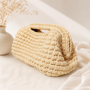 Pu&ograve; includere: Una pochette beige realizzata in materiale spesso intrecciato simile alla rafia. La borsa presenta una forma rettangolare arrotondata e strutturata con un'apertura superiore e un dettaglio a bordo intrecciato lungo la cornice, con trama a uncinetto.