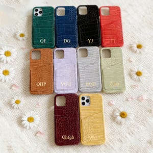 May include: A collection of ten colorful phone cases with a crocodile skin texture. The cases are in various colors, including green, blue, black, red, orange, lavender, gray, and yellow. Each case has a card slot and gold lettering. Daisies are scattered around the cases.