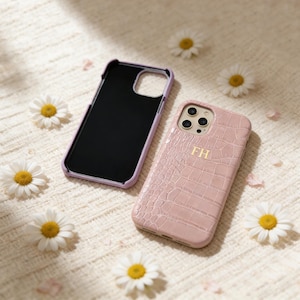 Puede incluir: Dos fundas de teléfono, una lavanda y otra rosa, descansan sobre una superficie crema con margaritas. La funda rosa tiene una textura de cocodrilo e iniciales doradas. La funda lavanda está abierta, revelando un interior negro.