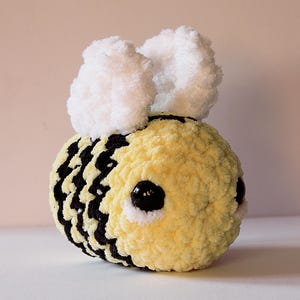 May include: A handmade, plush bee toy with a yellow body and black stripes. It features large, black eyes and fluffy white wings. The toy is crafted from soft, textured yarn.