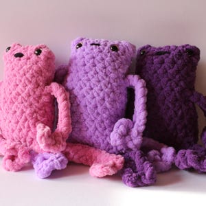 May include: Three handmade crochet frog plushies in shades of pink, purple, and dark purple. Each frog has a textured, fuzzy appearance with small black eyes and long limbs. The frogs are arranged together, showcasing their different colours.