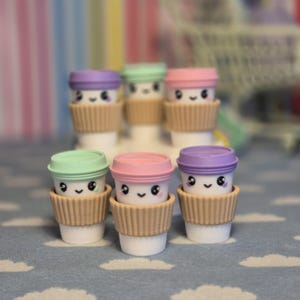 May include: Six miniature coffee cup figurines with smiling faces. Each cup has a different coloured lid: purple, green, and pink. The cups have a brown paper sleeve and are set on a blue surface with a cloud pattern.